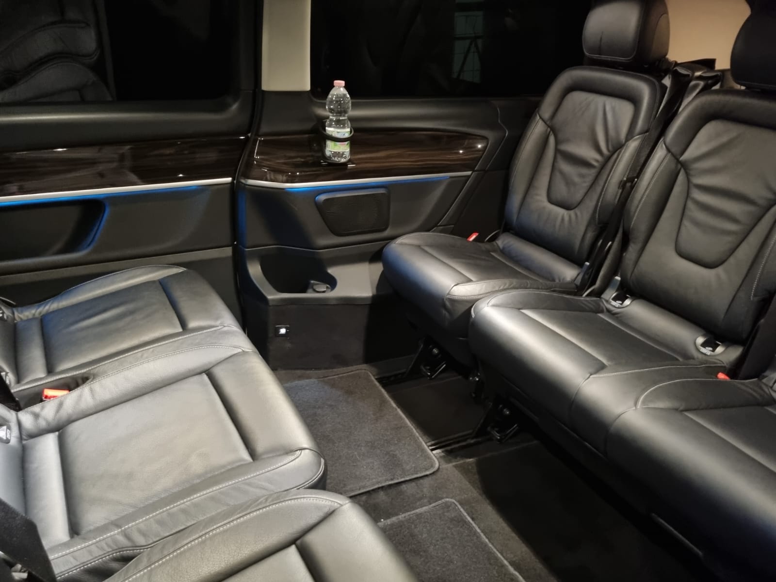 Interior of a Mercedes-Benz V-Class luxury van showing black leather vis-à-vis seating, a folding table, and ambient lighting, configured for business or group travel