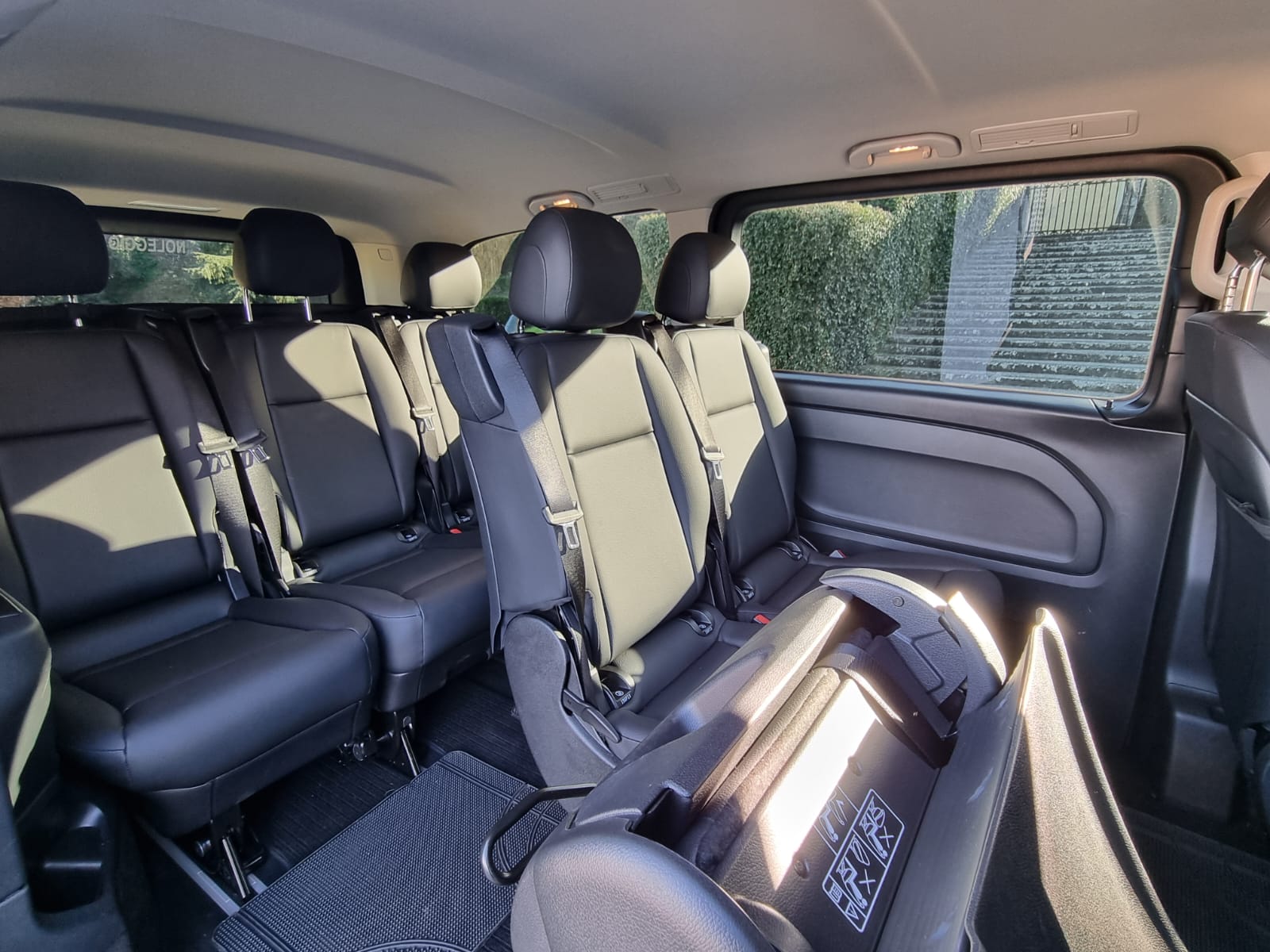 View from the front seats towards the back of a Mercedes luxury van, showing two full rows of forward-facing passenger seats with headrests, highlighting passenger capacity.