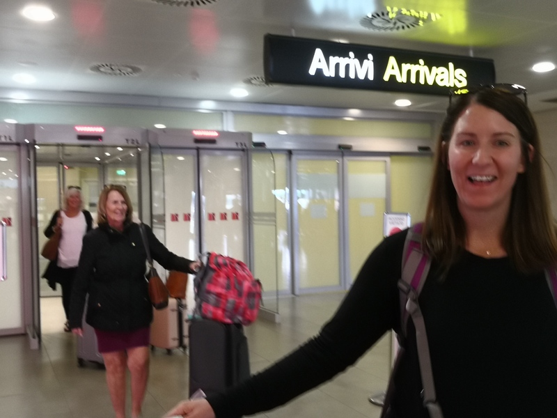 Two happy travelers with luggage arriving at an airport gate, greeted by a Florence Pisa Taxi driver for a stress-free transfer