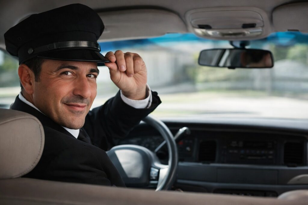 Professional chauffeur in uniform turning to greet from the driver's seat, offering friendly and reliable taxi service in Tuscany