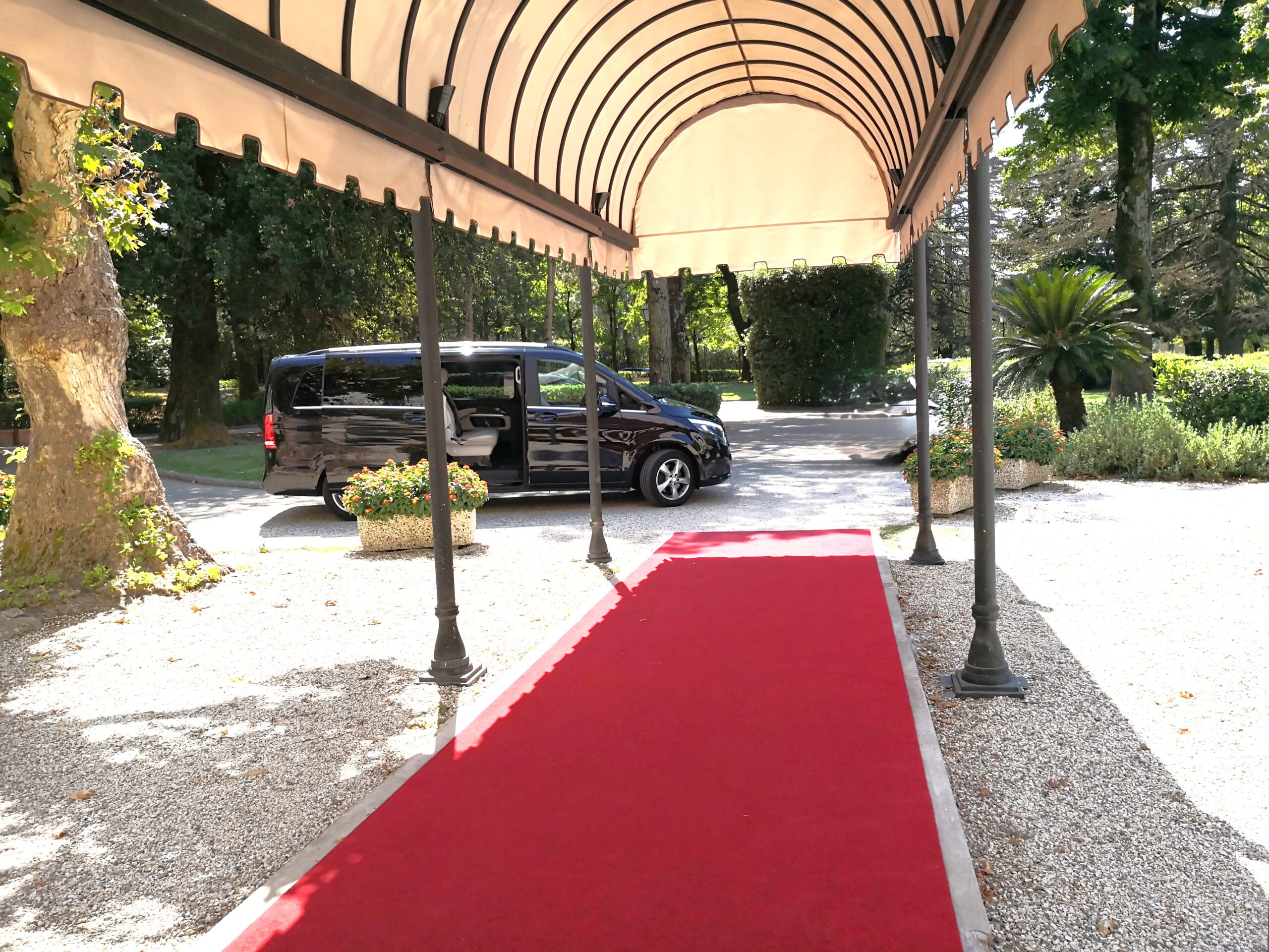 A black Mercedes-Benz V-Class luxury van awaits with an open door and a red carpet at the entrance of the Grotta Giusti Spa Resort in Tuscany on a sunny day.